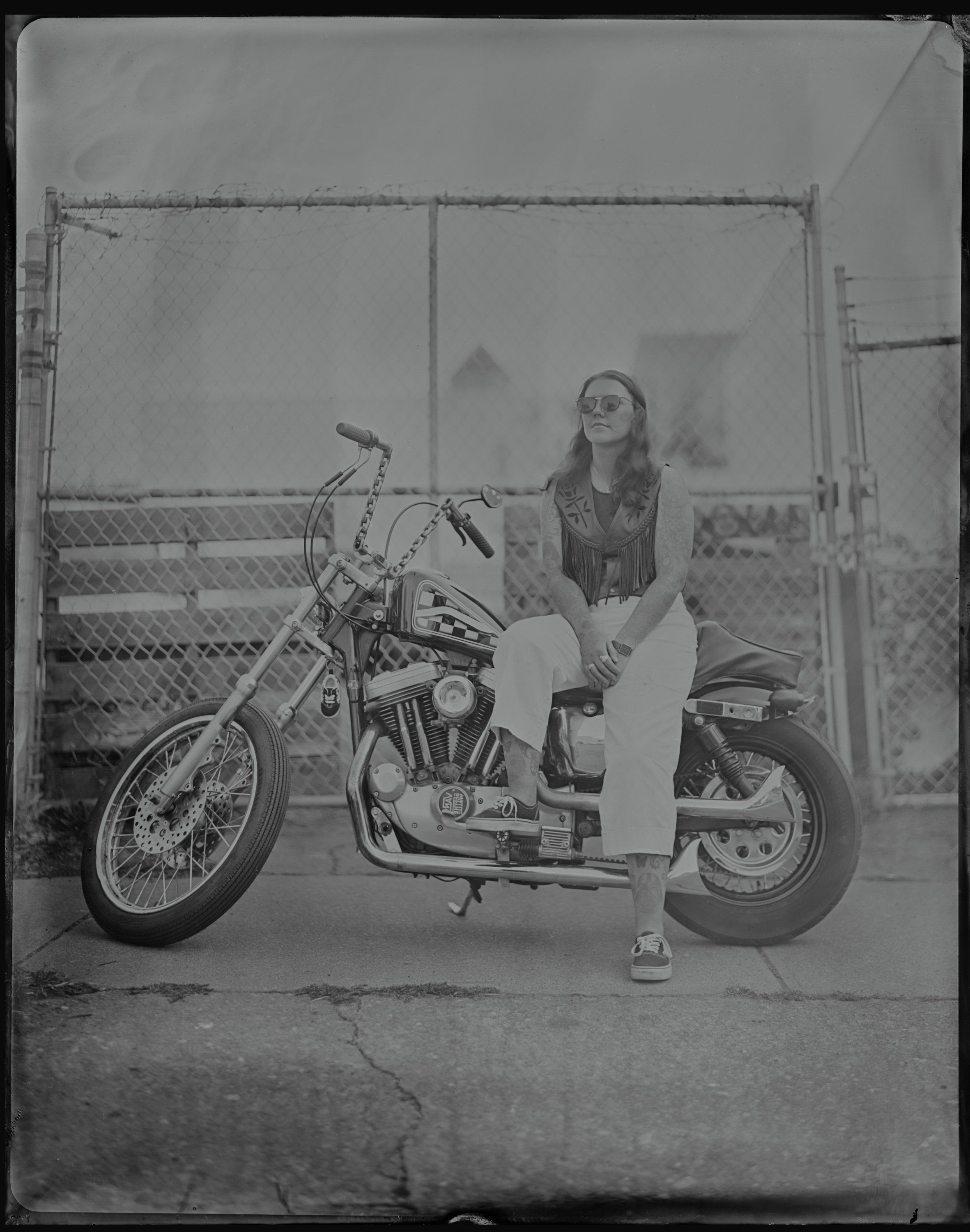 Woman in sunglasses and light pants sits sidesaddle on a vintage motorcycle against a chain-link fence in this black and white tintype