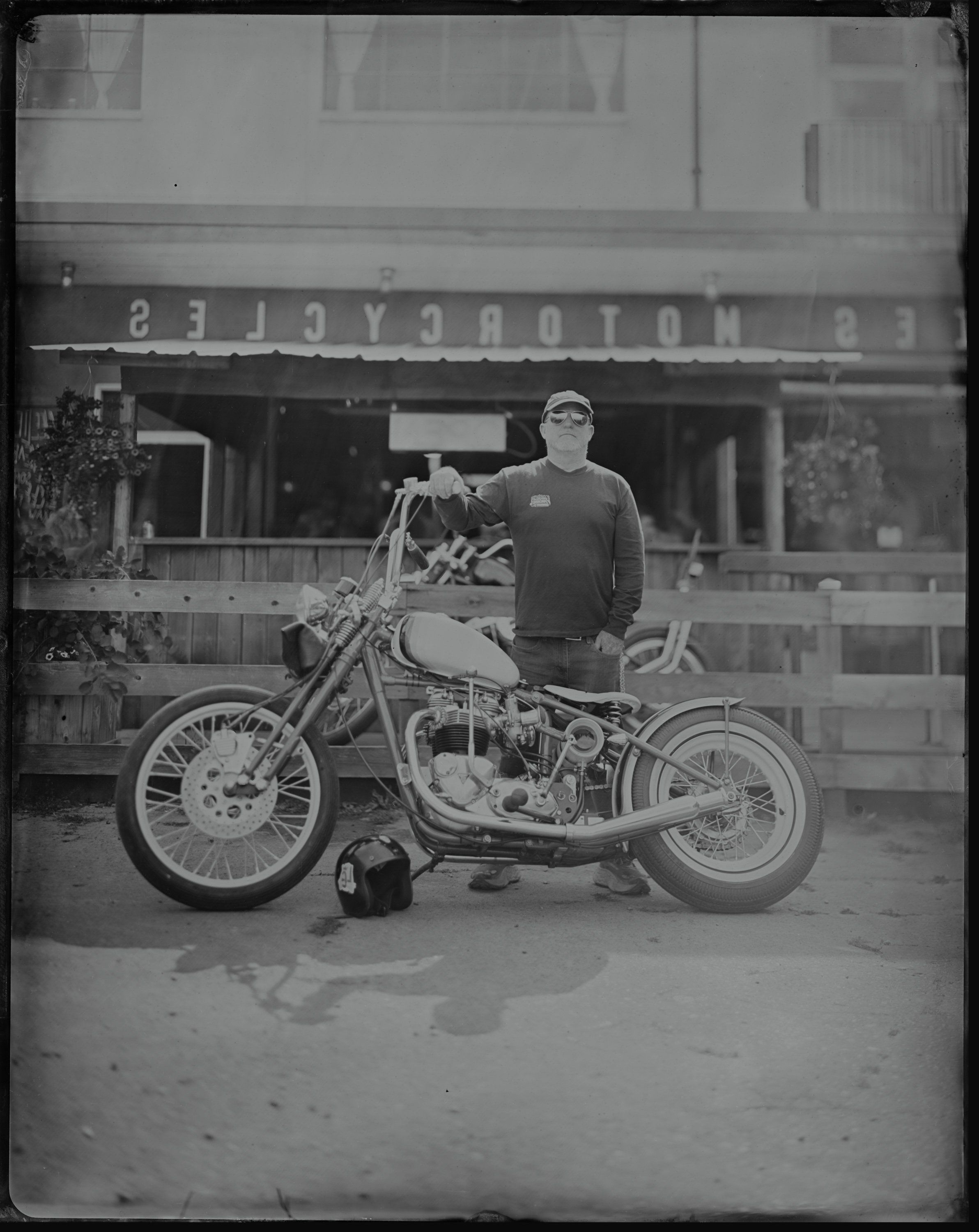 Man in glasses posing with vintage motorcycle in front of storefront, black and white photograph with soft natural light