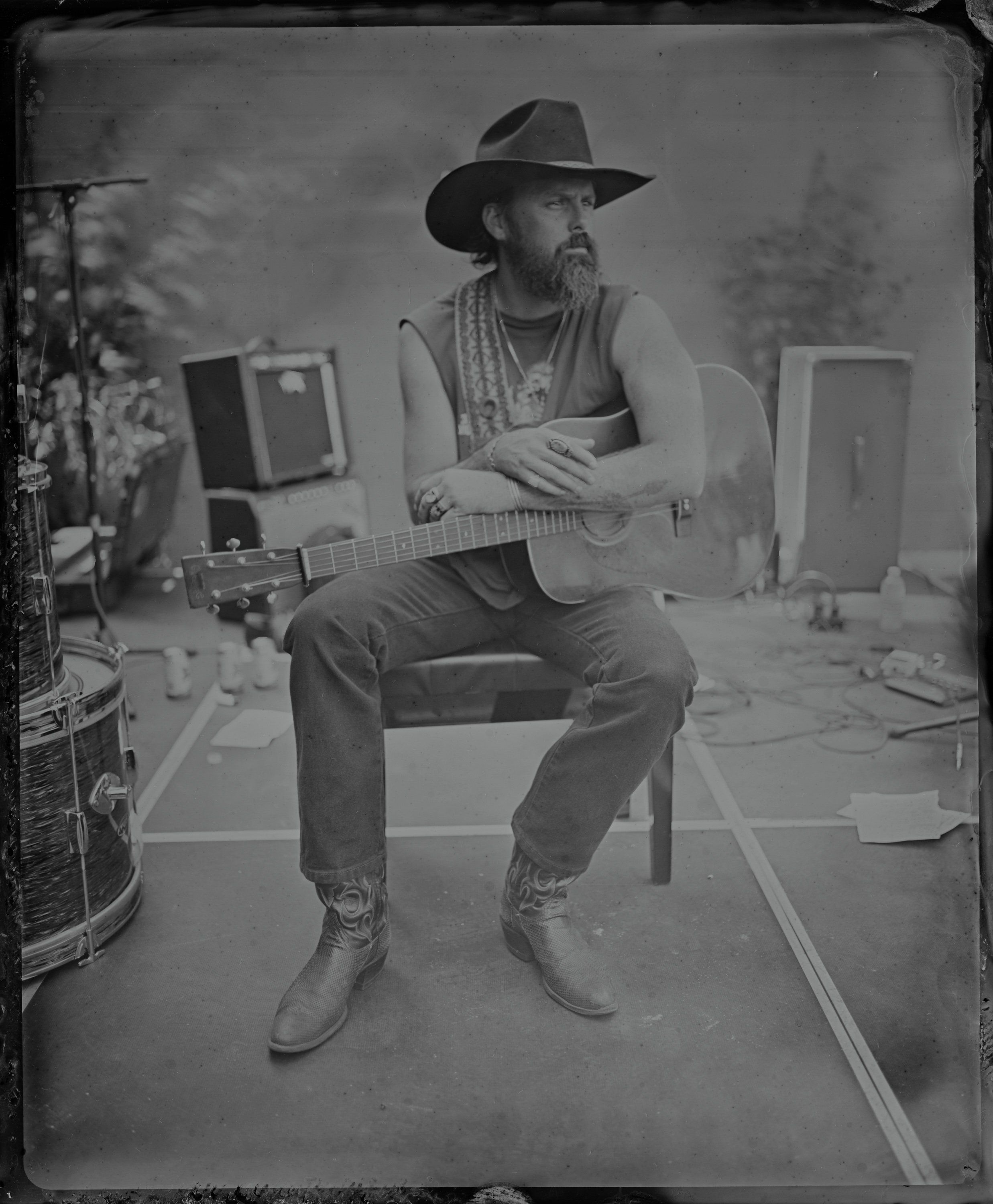 Bearded musician in cowboy hat sits with acoustic guitar in a parking lot studio setting, surrounded by amplifiers and studio equipment, rendered in tintype…