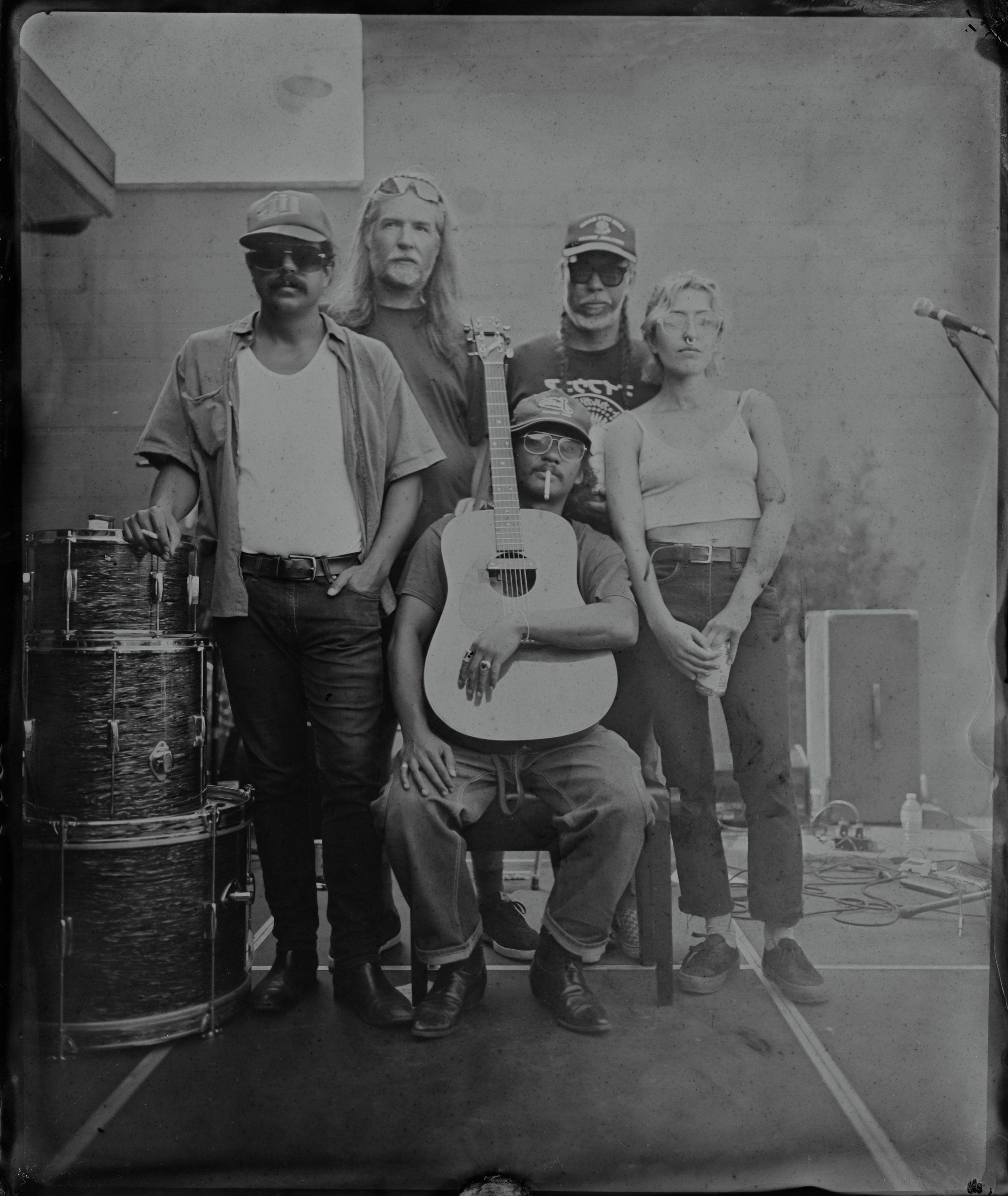 Four musicians pose indoors with drum kit and acoustic guitar, rendered in silver and tin tintype tones with natural side lighting