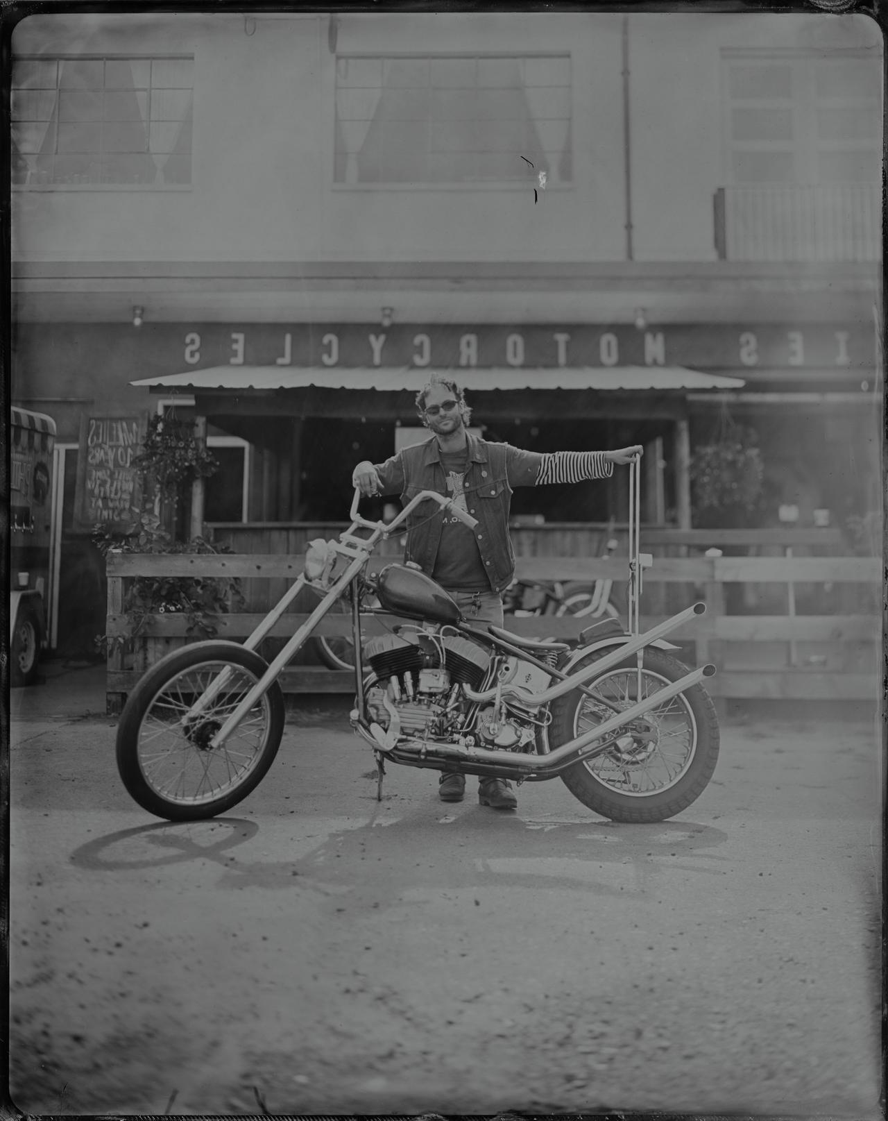 Man in sunglasses and denim vest leans against motorcycle at Wheelies parking lot fest, black and white tintype