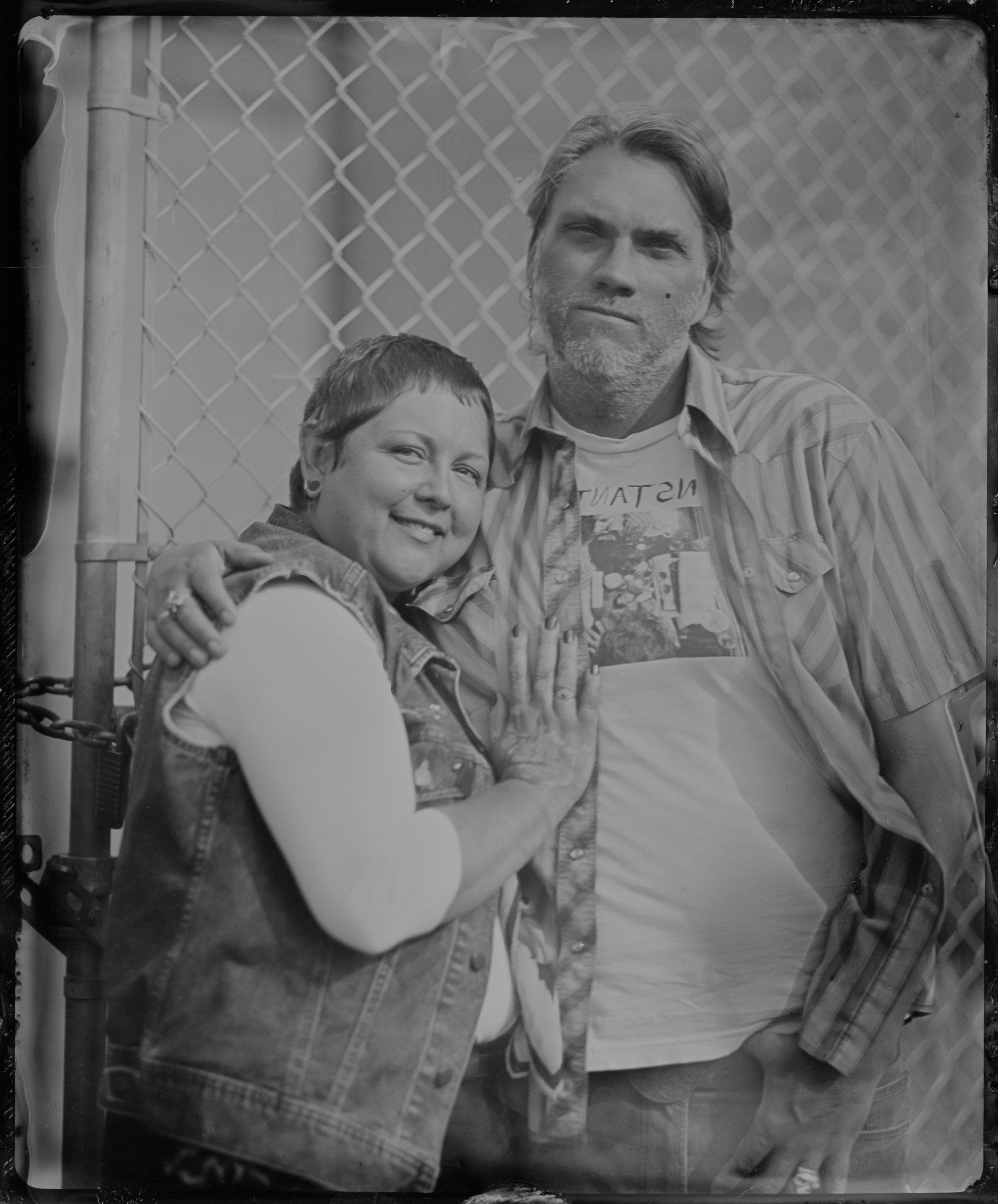 Man and young boy in denim, smiling at camera against chain-link fence backdrop, tintype style black and white portrait