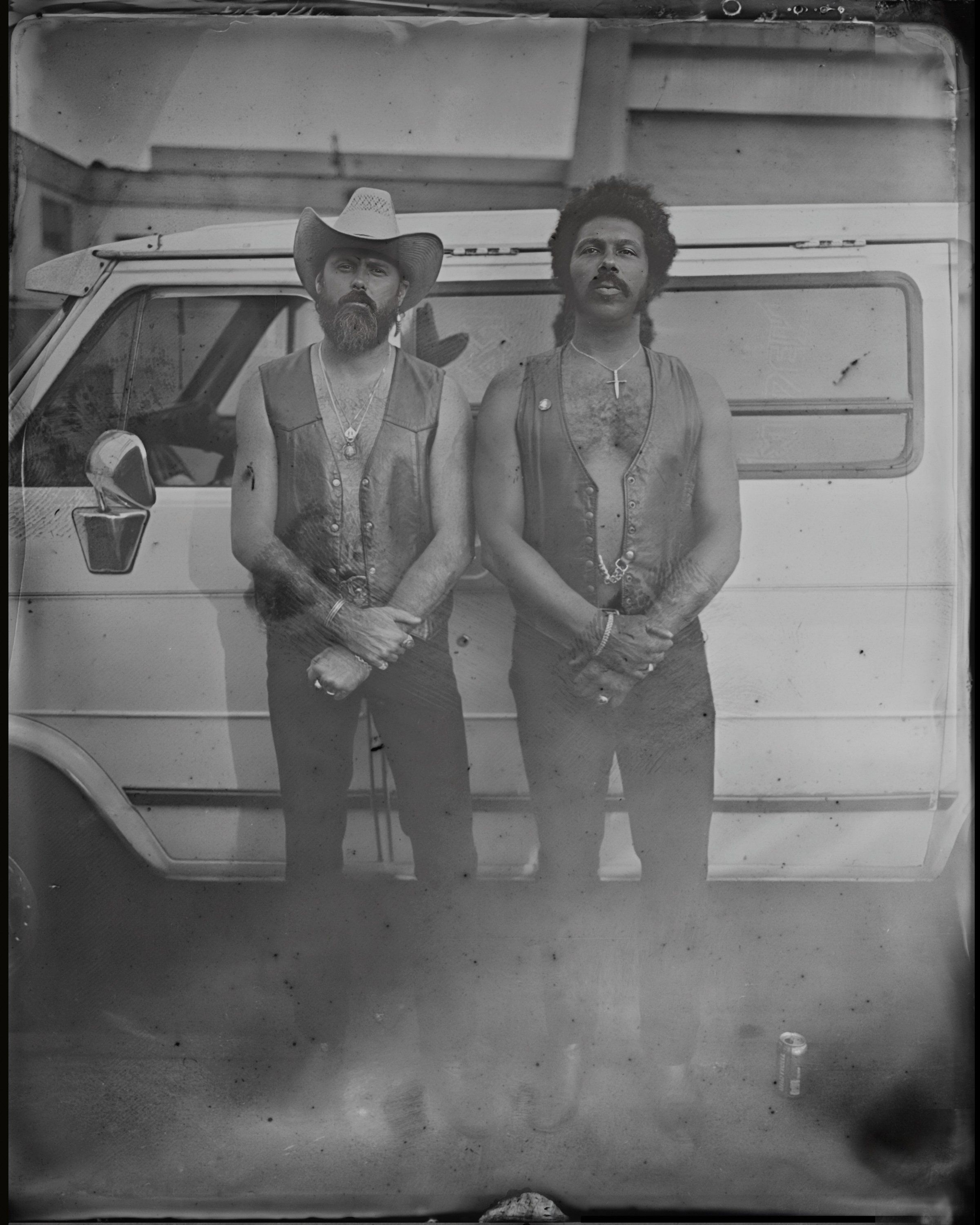 Two mustachioed men in cowboy hats stand before a silver trailer, wearing open vests and long beaded necklaces in this tintype-style black and white portrait
