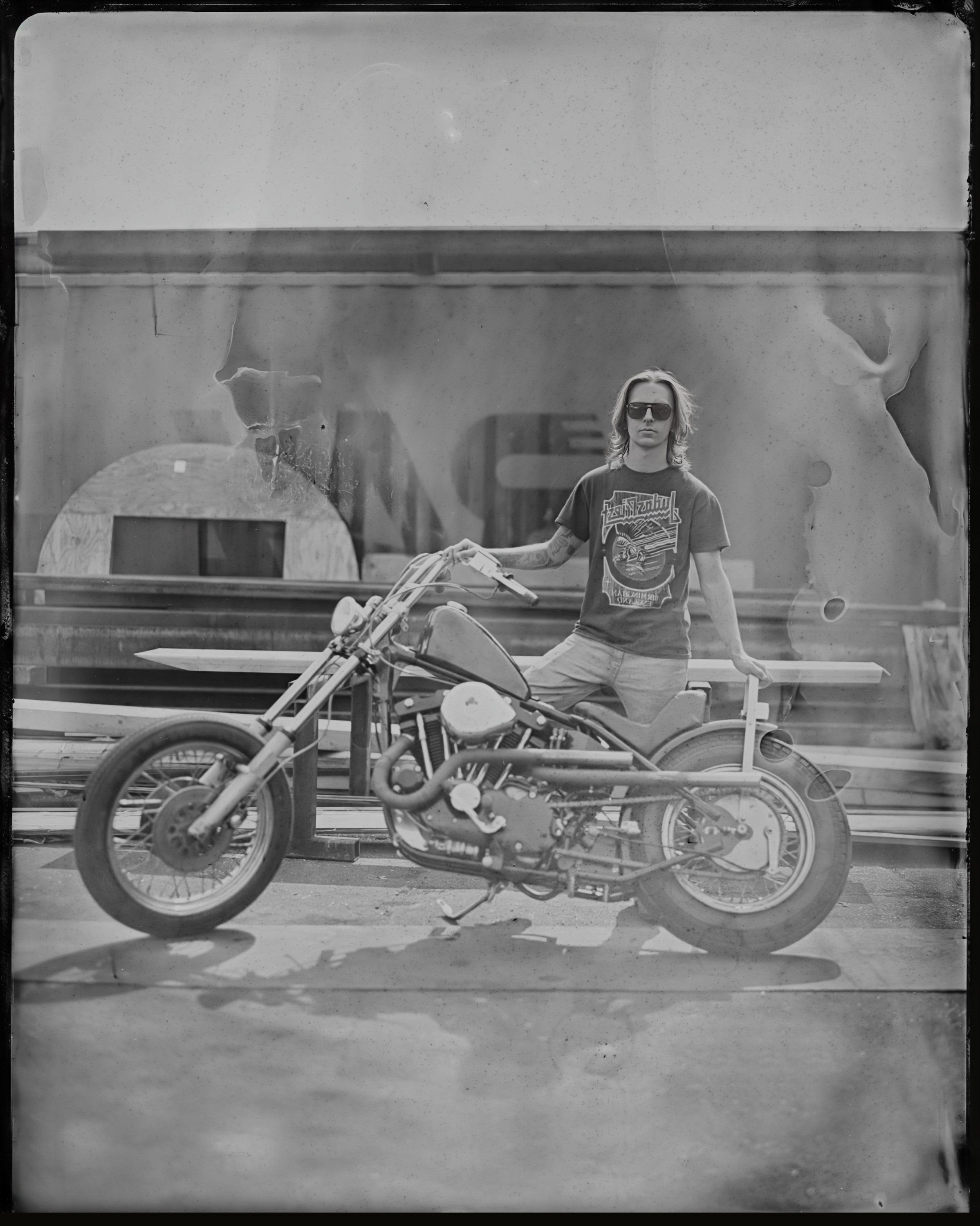 Person in graphic tee sitting on custom motorcycle in parking lot, captured as tintype with vintage aesthetic and soft natural light