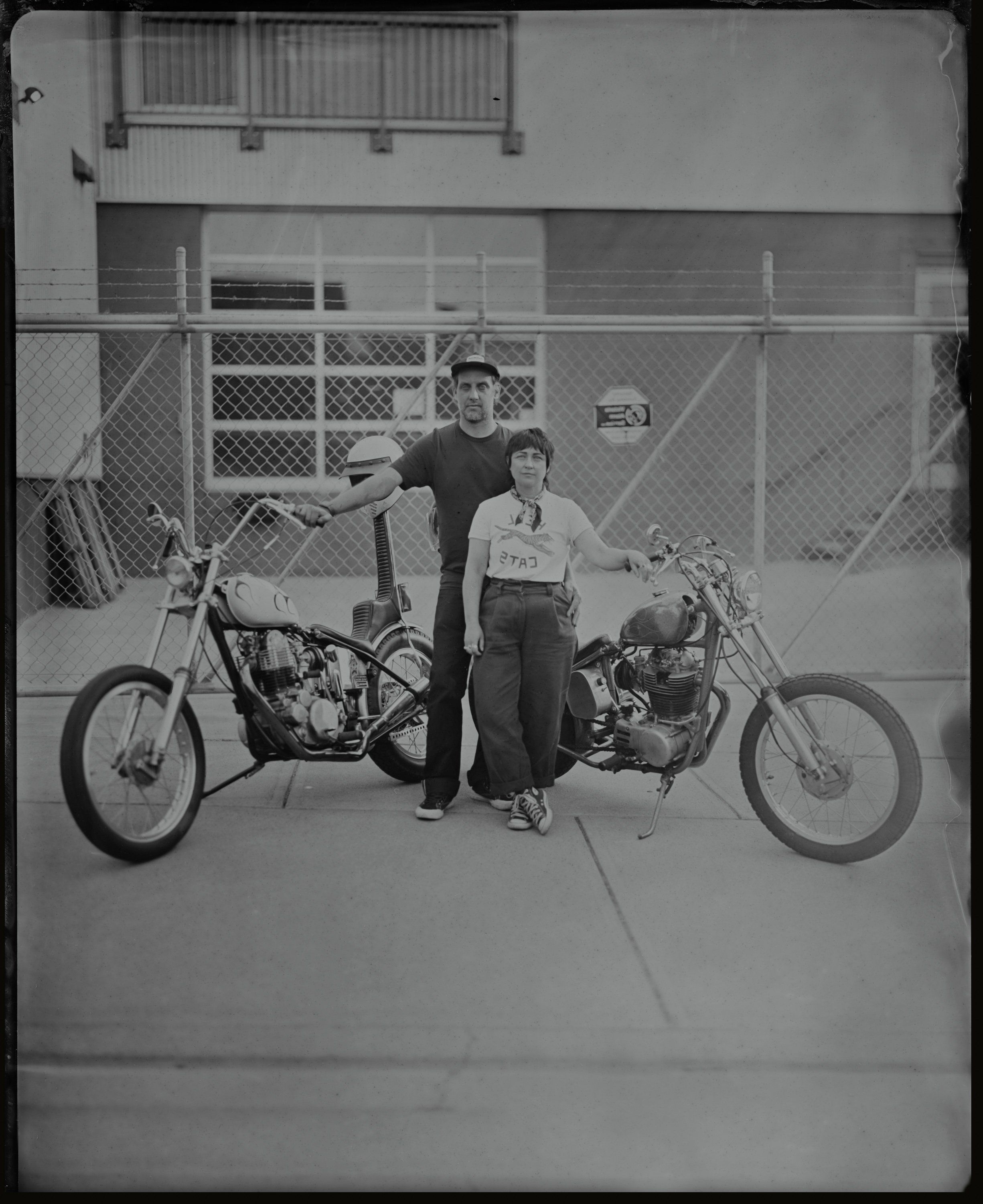 Two people pose between custom choppers in a parking lot, framed by chain-link fence and industrial building, rendered in stark black and white