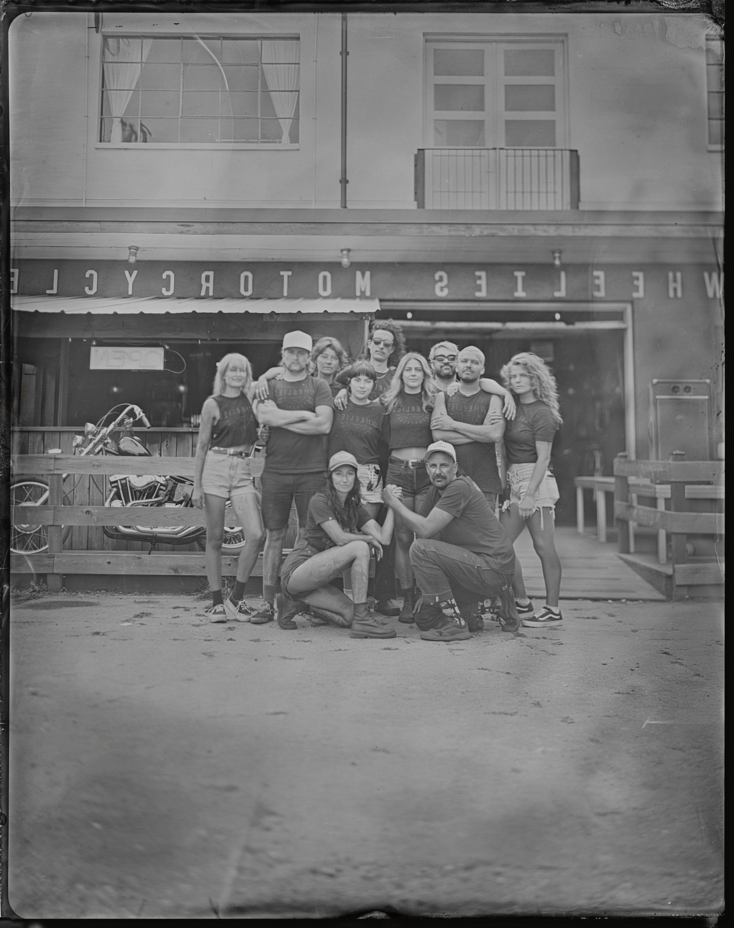 Group portrait of approximately fifteen people posing together in a parking lot outside a storefront, rendered in black and white tintype style