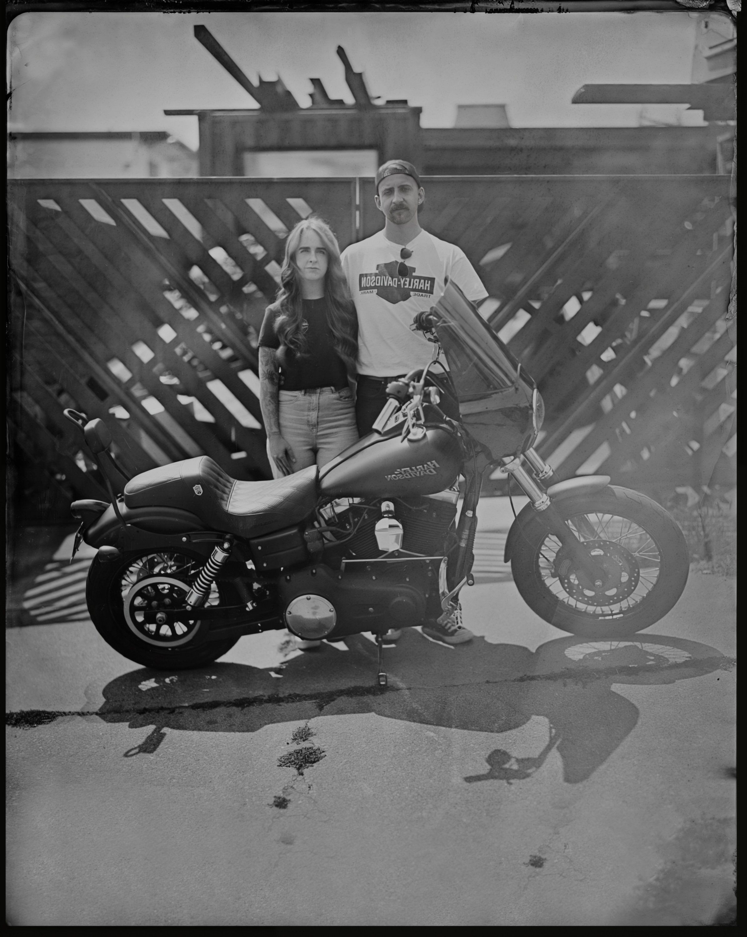 Couple posed beside a motorcycle against industrial backdrop with geometric shadow patterns, black and white tintype style