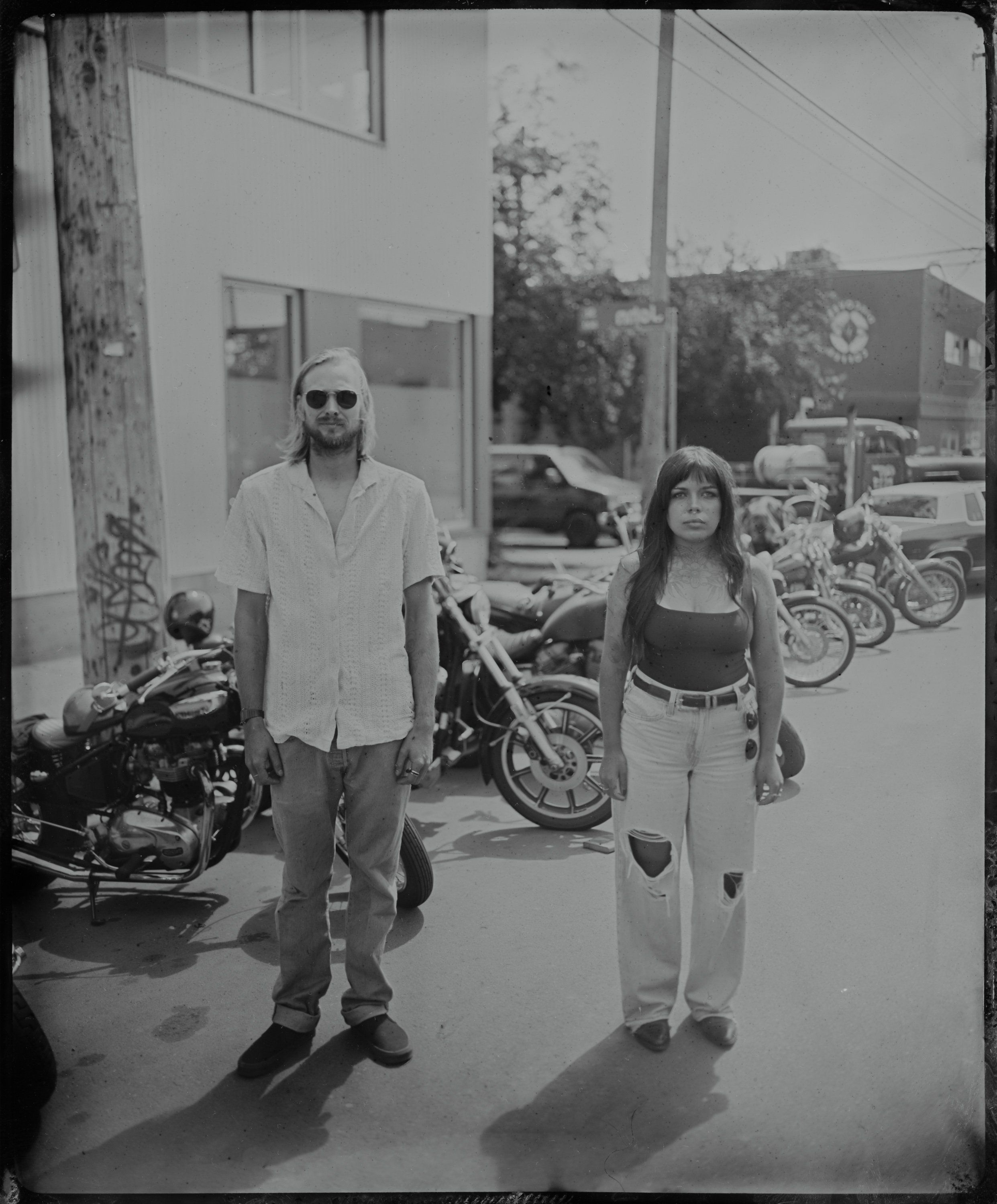 Man in sunglasses and woman walking hand-in-hand past motorcycles in a parking lot, bright sunlight casting sharp shadows