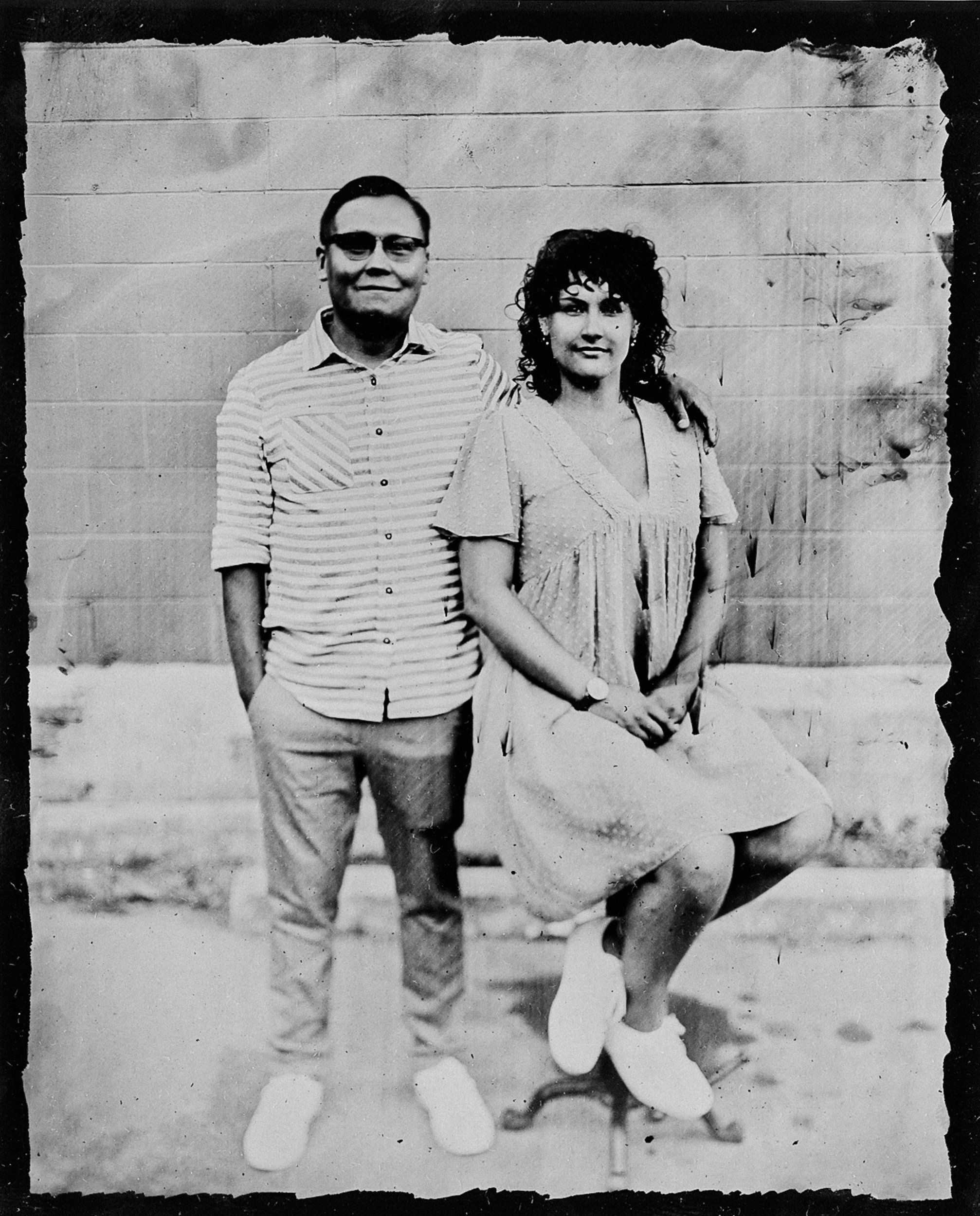 Couple in casual 1970s attire smiling at camera against weathered concrete wall, tintype effect with vintage grain and distressing
