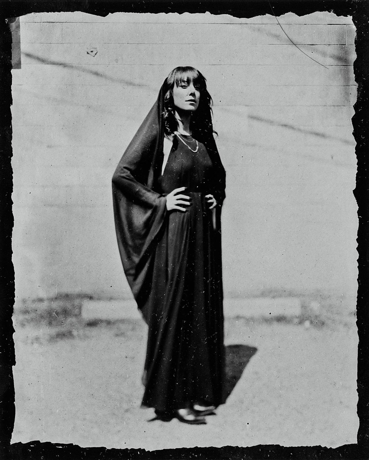 Woman in long dark dress with shawl and pearl necklace stands against textured wall, gazing upward in contemplative pose