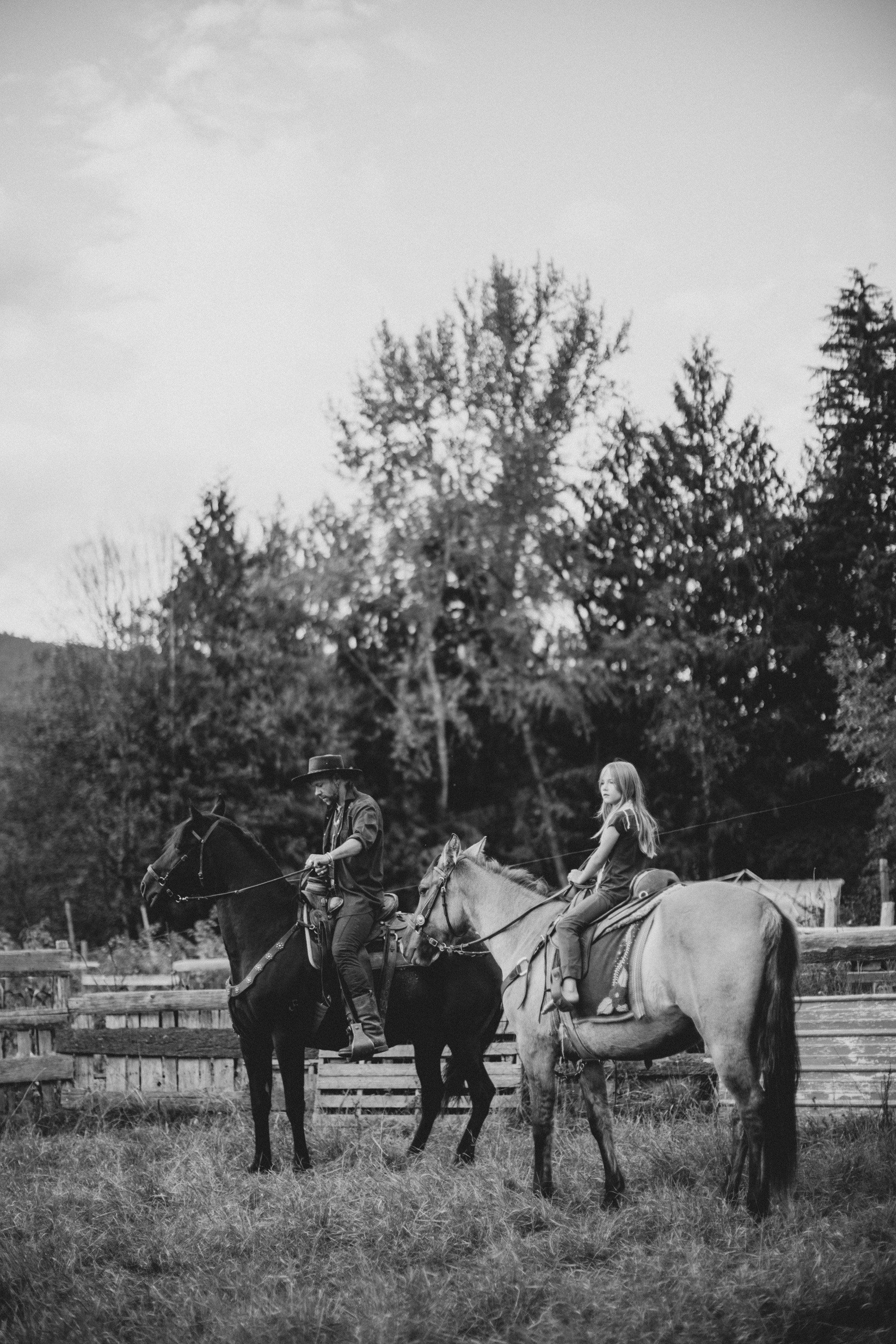 Two riders on horseback in a pastoral field with wooden fencing and forested hills behind them in soft, overcast light