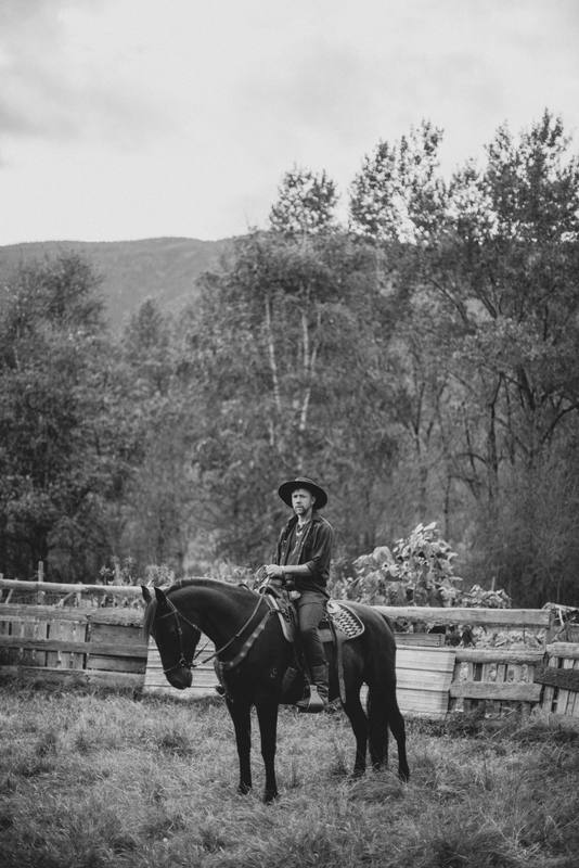 Cowboy on dark horse stands in ranch pasture with wooden fence and forested hills under overcast sky