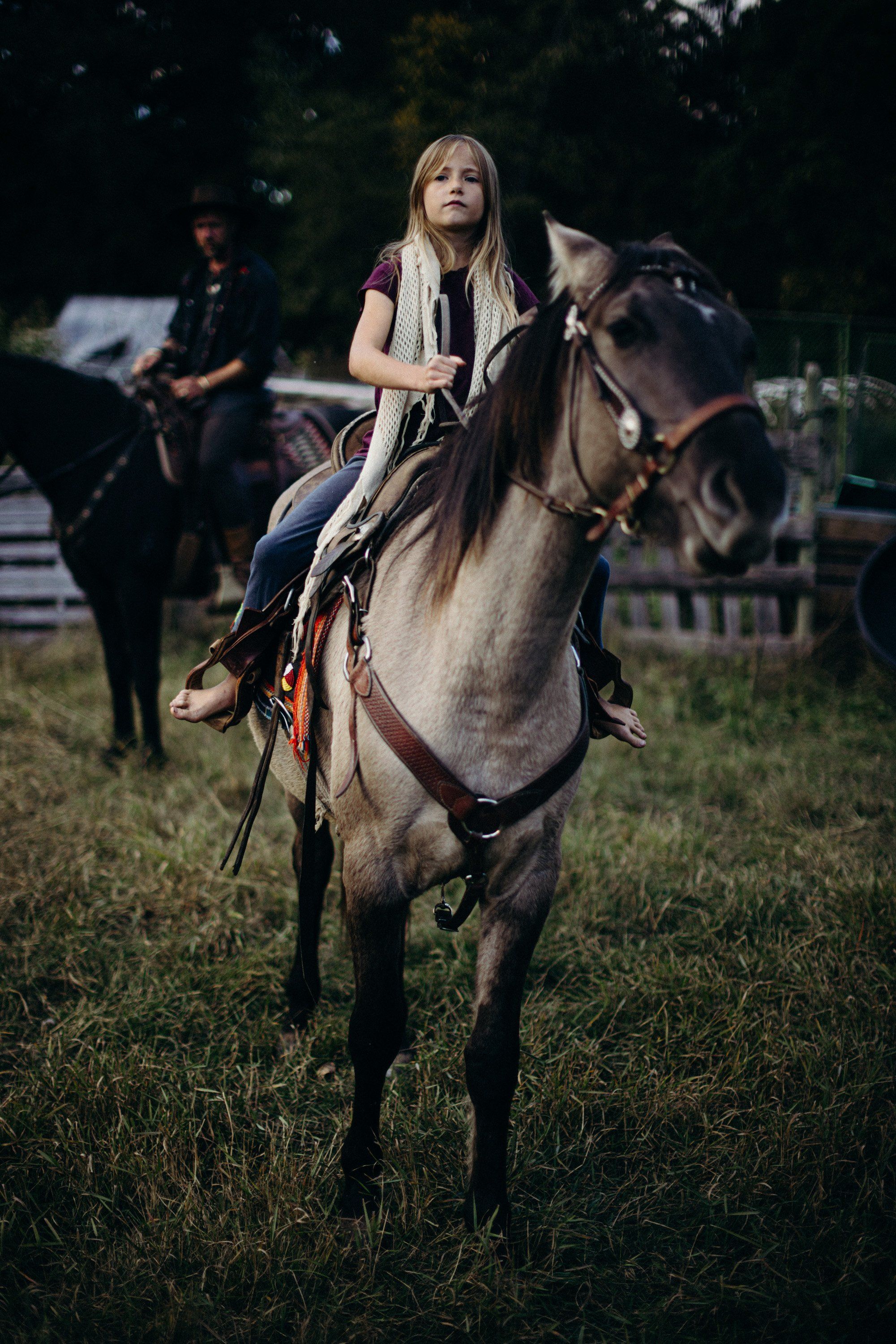 Woman with long blonde hair riding a gray horse across a grassy field at dusk, wearing a cream linen shirt and leather saddle