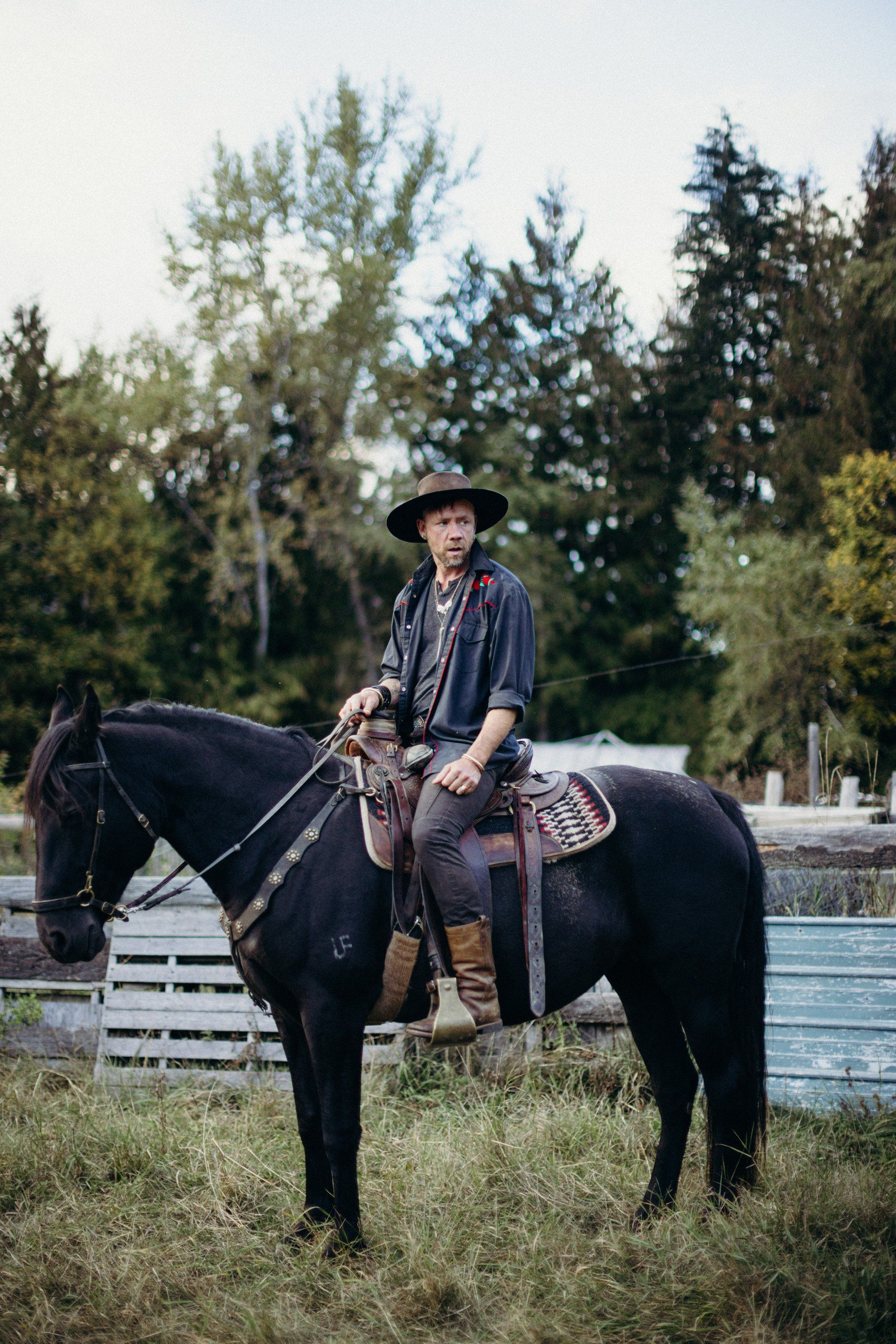 Cowboy in denim and hat on black horse in grassy pasture with white fencing and evergreen trees under soft afternoon light