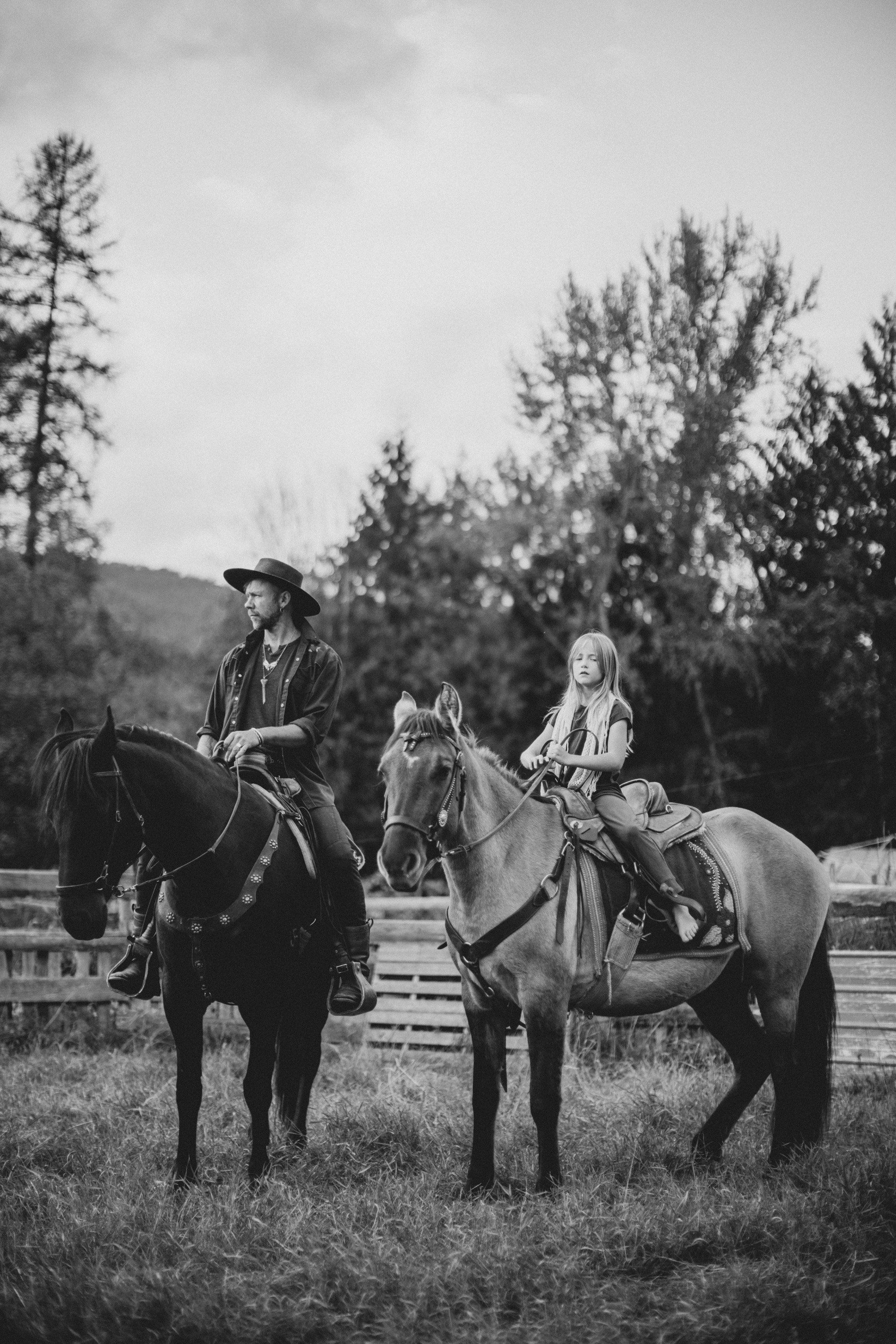 Two riders on horseback in a pastoral field with evergreen trees, rendered in soft black and white tones