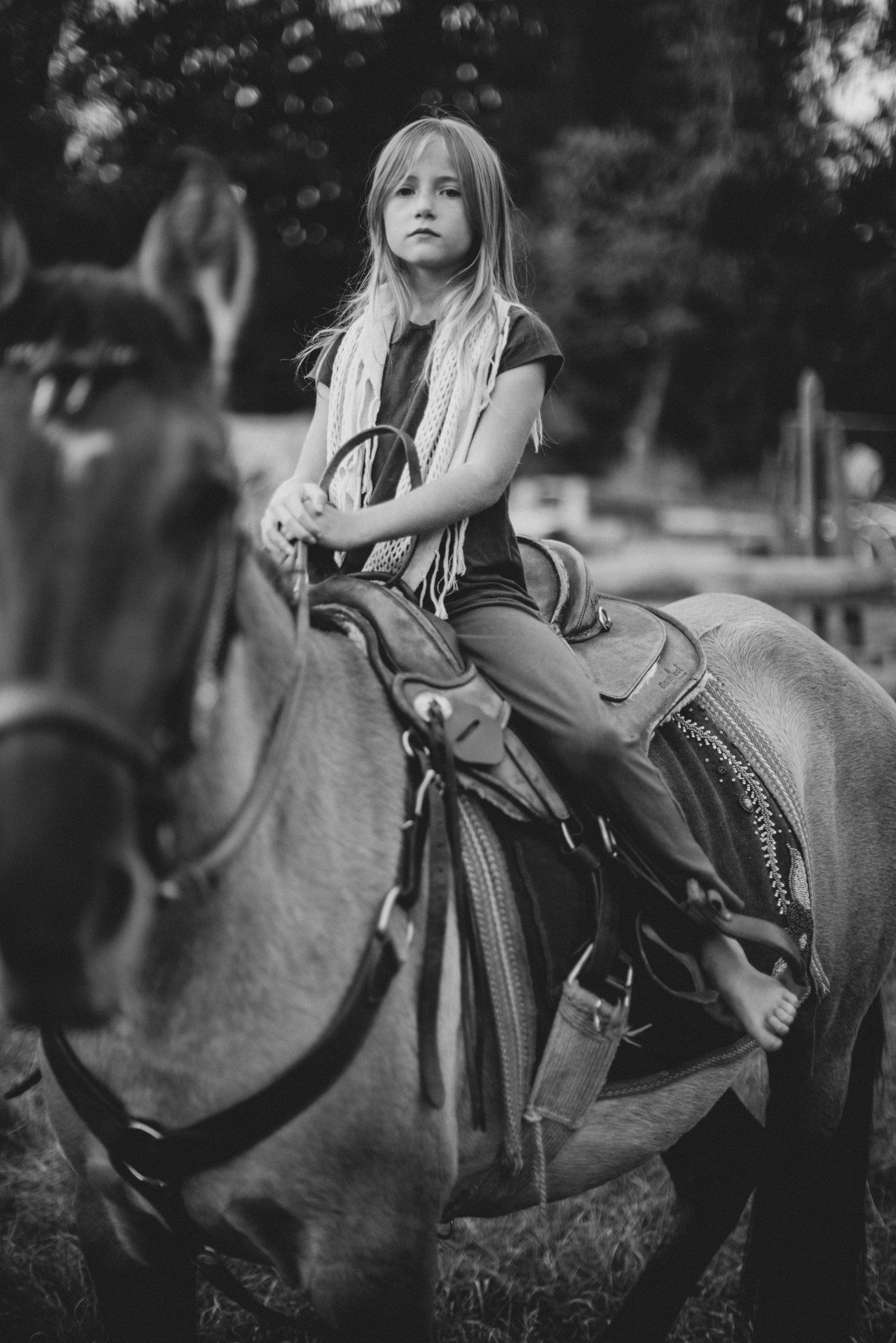 Young woman on horseback in black and white, long blonde hair, contemplative gaze, dappled forest light behind