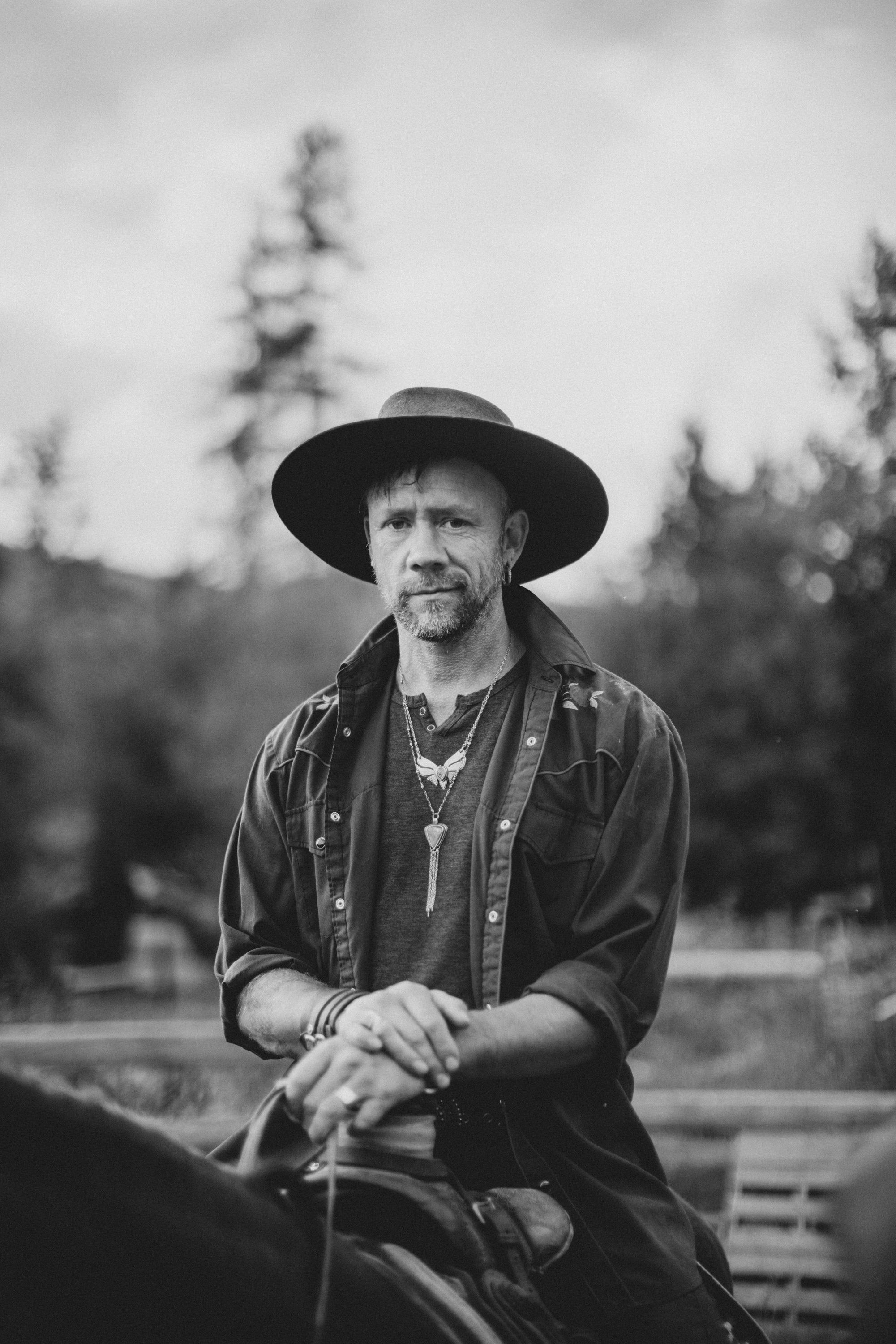 Bearded man in wide-brimmed hat and layered shirt sits contemplatively against forested backdrop in black and white portrait