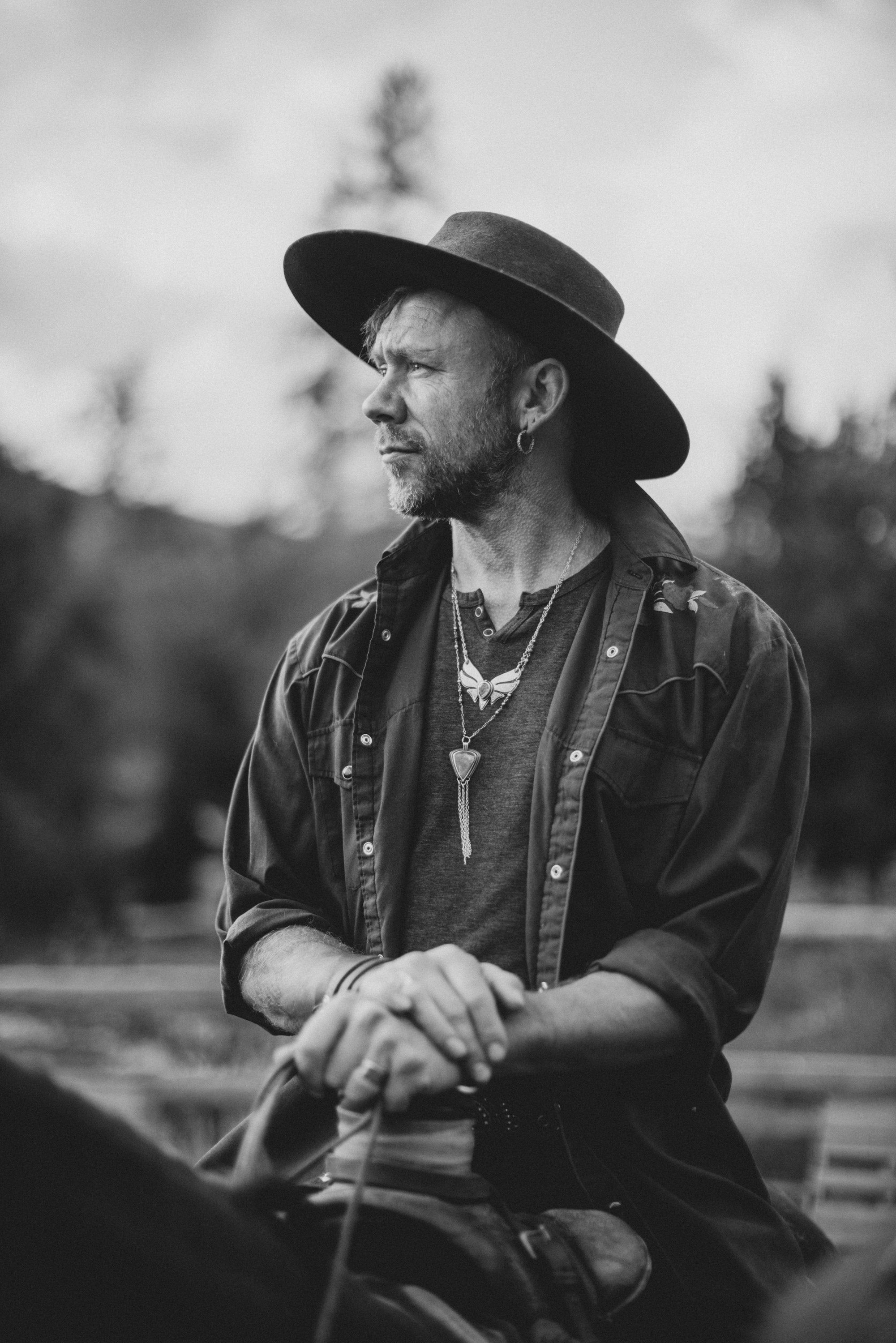 Man in black cowboy hat and embroidered shirt gazes thoughtfully into distance, forested landscape soft-focused behind him in moody black and white