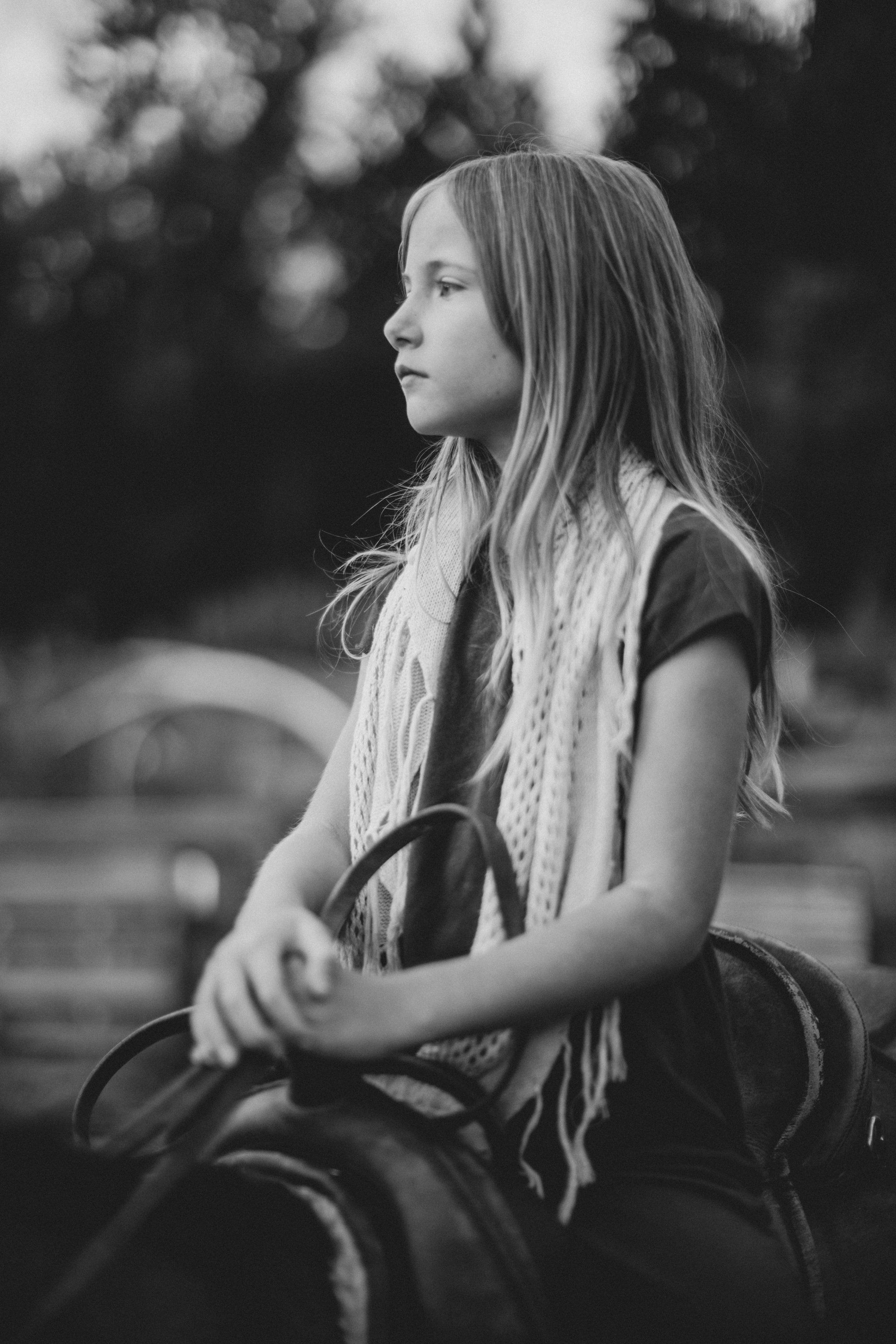 Young girl in profile sitting on a chair, long hair catching light, wearing a scarf, soft-focus natural setting in black and white