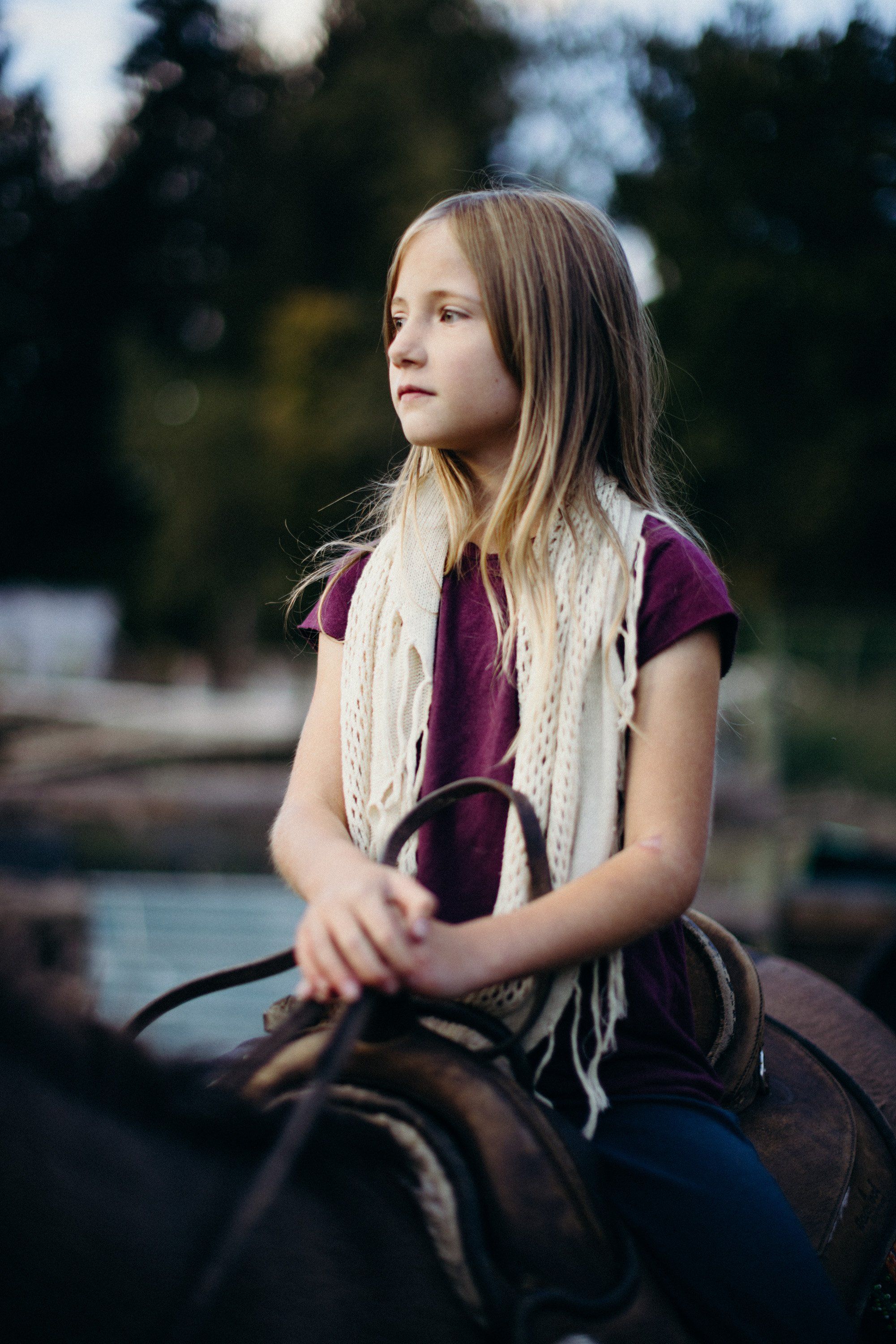 Young girl in burgundy shirt and cream scarf sits on saddle, gazing thoughtfully across ranch setting with evergreen trees