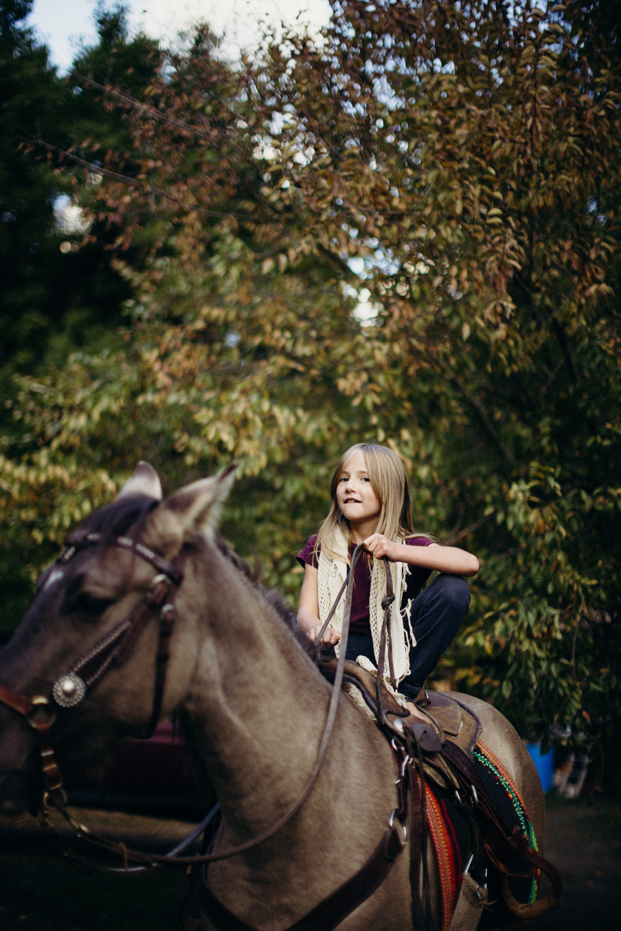 Young woman in vintage scarf sits on brown horse beneath golden autumn foliage, looking upward with gentle expression