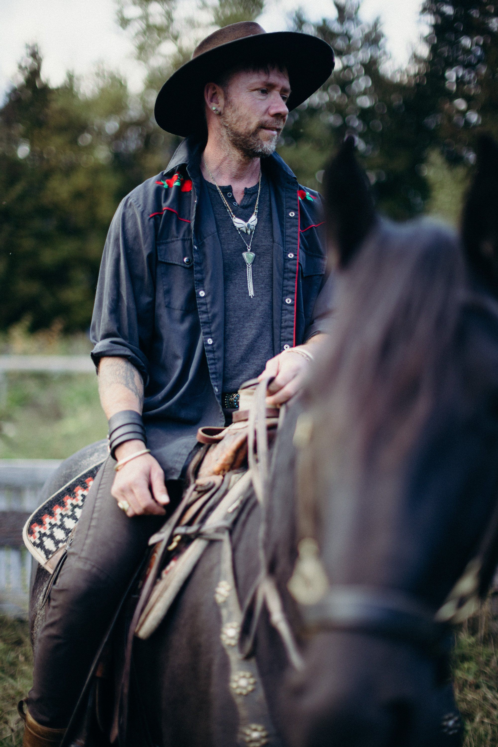 Man in black cowboy hat and denim shirt sits on horse, holding reins, with blurred figure in foreground and evergreen trees behind
