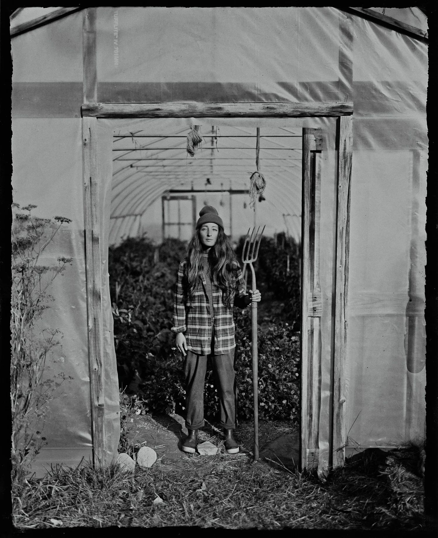 Woman in plaid shirt and beanie holding a pitchfork in a misty greenhouse, hung herbs overhead, contemplative studio portrait